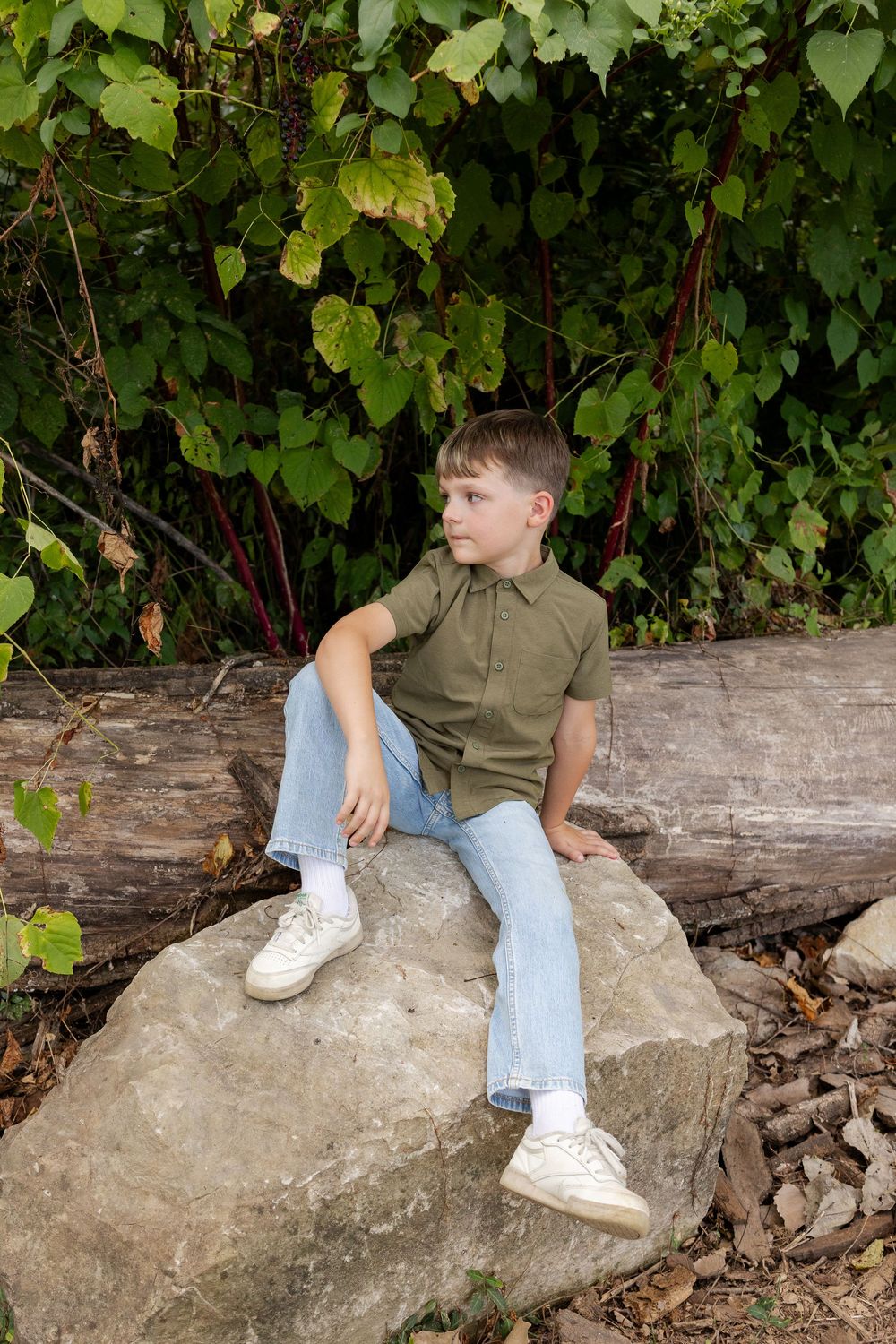 Young boy in an olive green button-up shirt and light jeans sits on a large boulder beside a fallen log and green leafy vines.