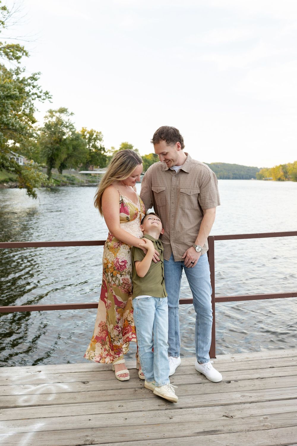 Smiling family of three embracing on a wooden dock beside a calm lake, with trees and a soft sky in the background.