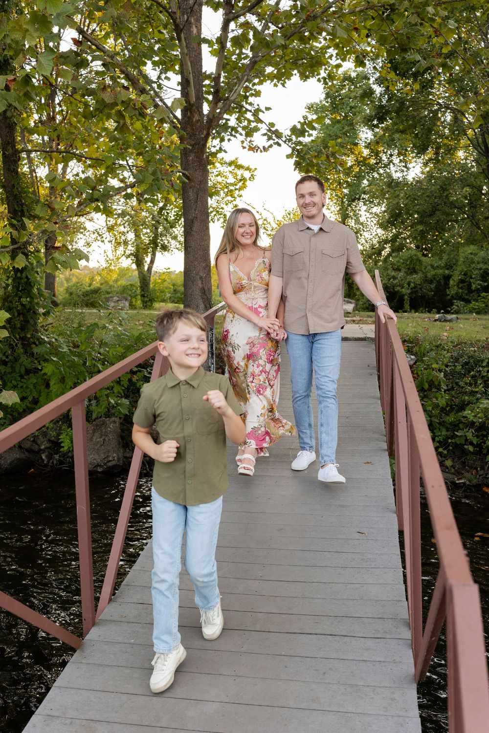 A smiling boy runs across a wooden footbridge while his parents walk hand in hand behind him in a leafy green park.