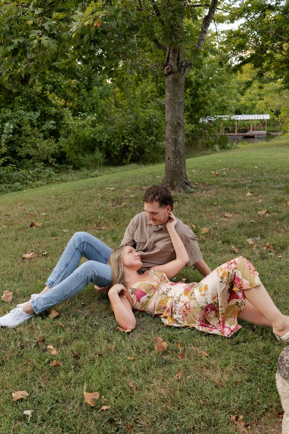 Couple relaxing on a grassy lawn under a shady tree, the woman in a floral dress resting her head on the man's lap in a park.