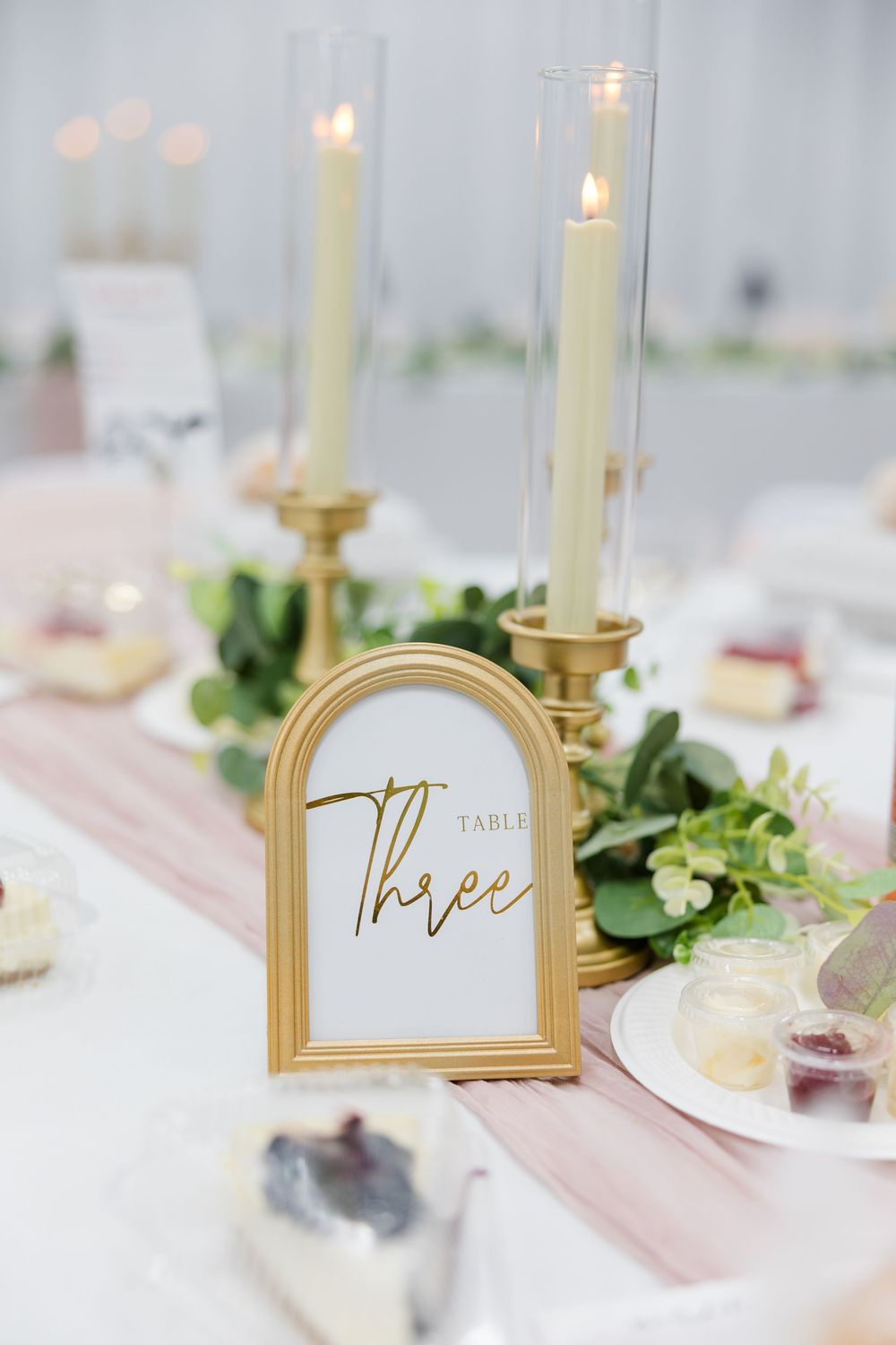 Gold-framed Table Three sign at elegant wedding reception with candlesticks and greenery centerpiece.