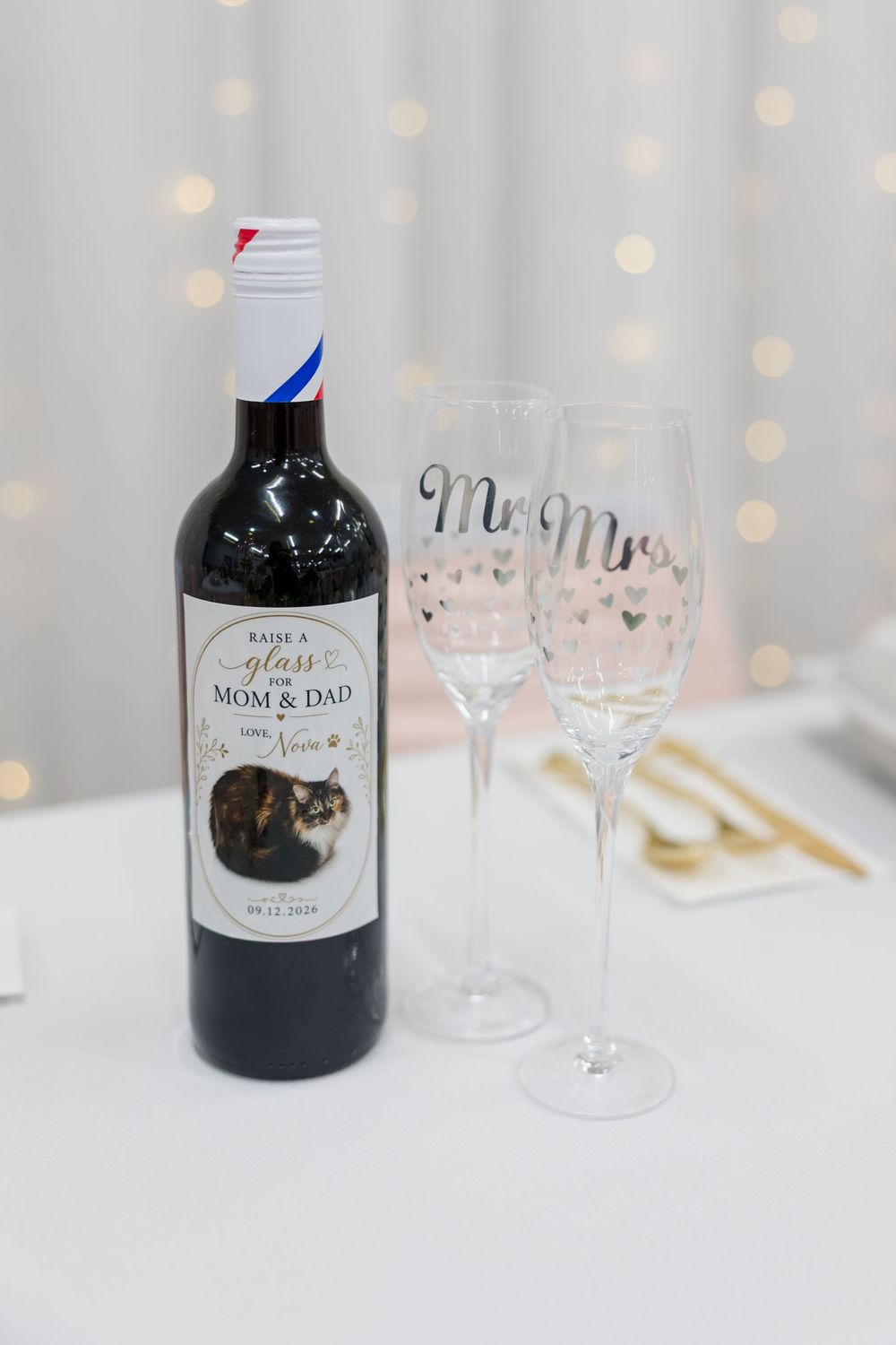 Custom wedding wine bottle labeled Mom & Dad beside Mr. and Mrs. champagne flutes on a white table.
