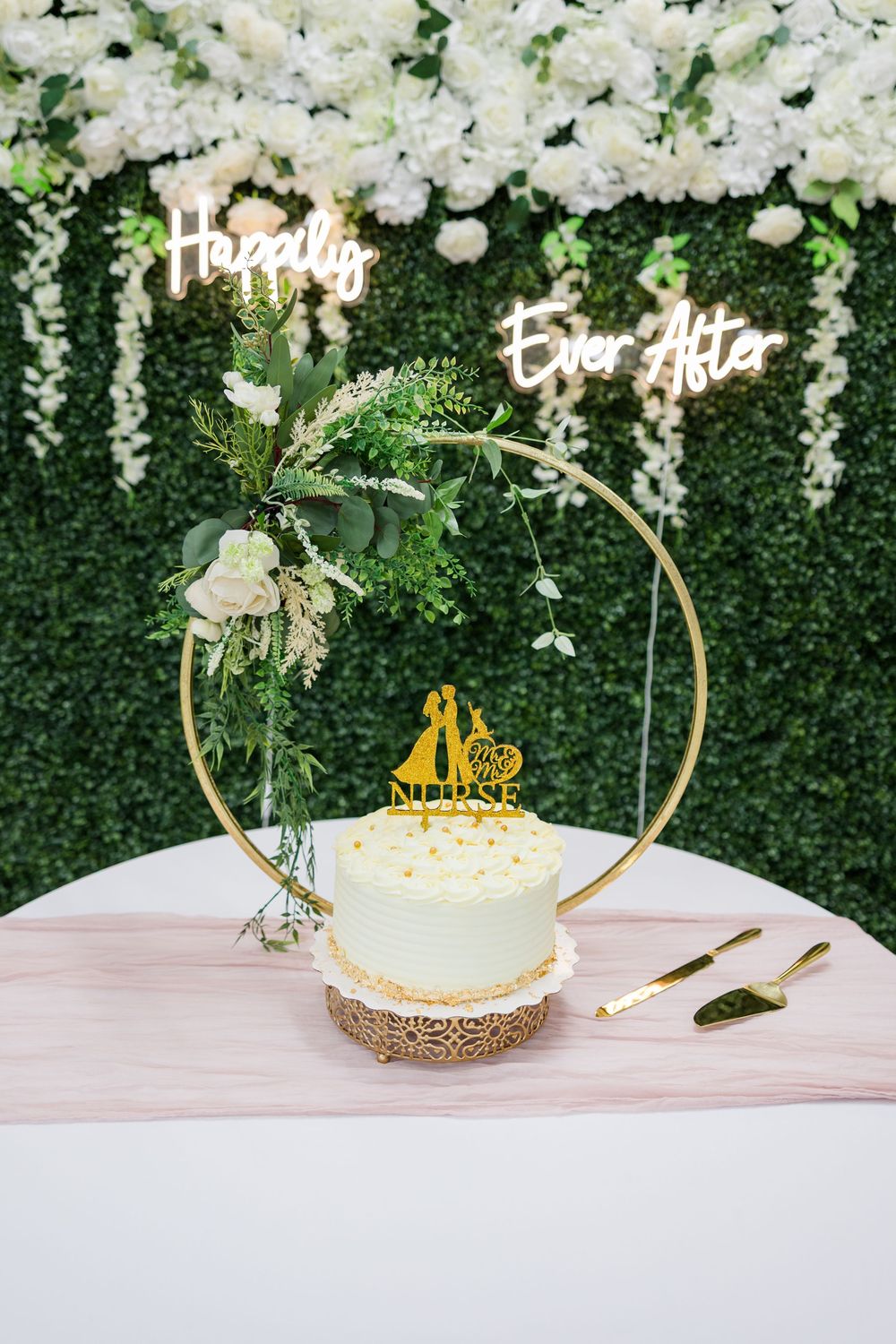 Elegant wedding cake with gold hoop stand, floral decor, and Happily Ever After neon sign on green wall backdrop.