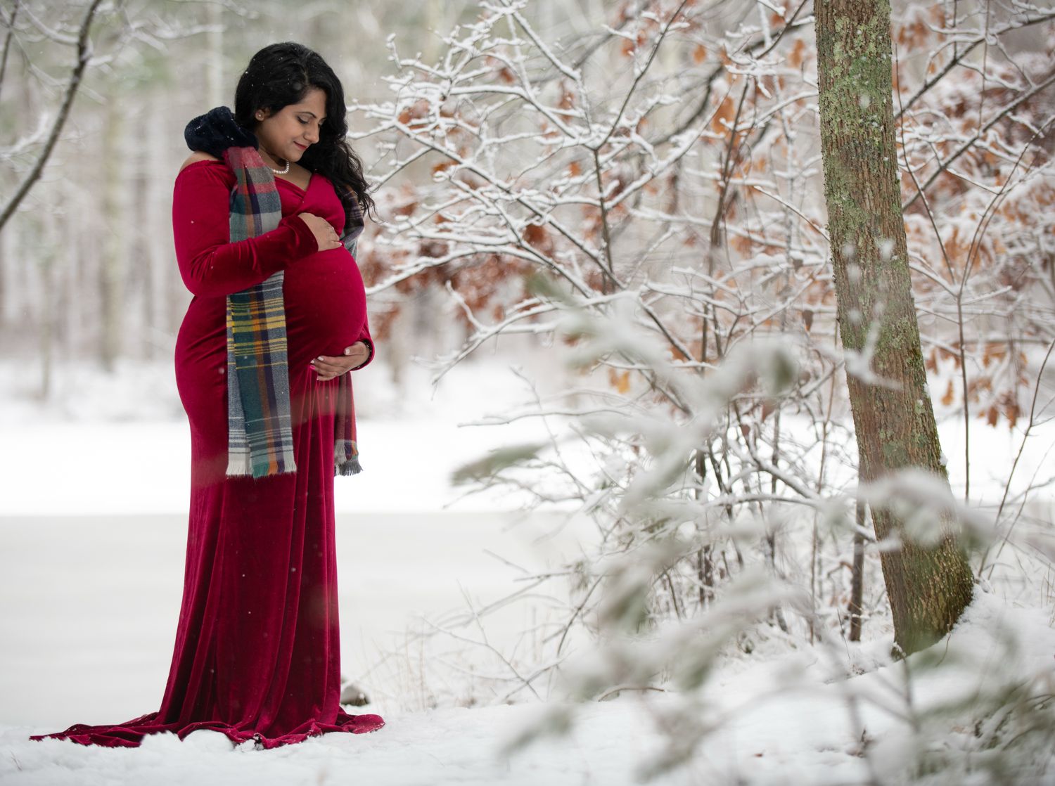 Pregnant woman in a flowing red maternity gown and plaid scarf cradles her baby bump in a snowy winter forest.