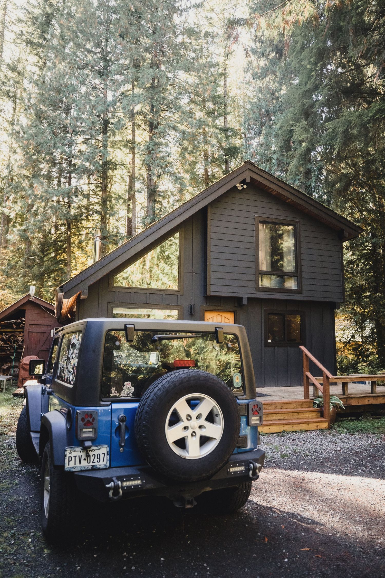 Blue Jeep Wrangler parked on gravel beside a modern dark-gray A-frame cabin with a wooden deck, surrounded by tall evergreen forest trees.