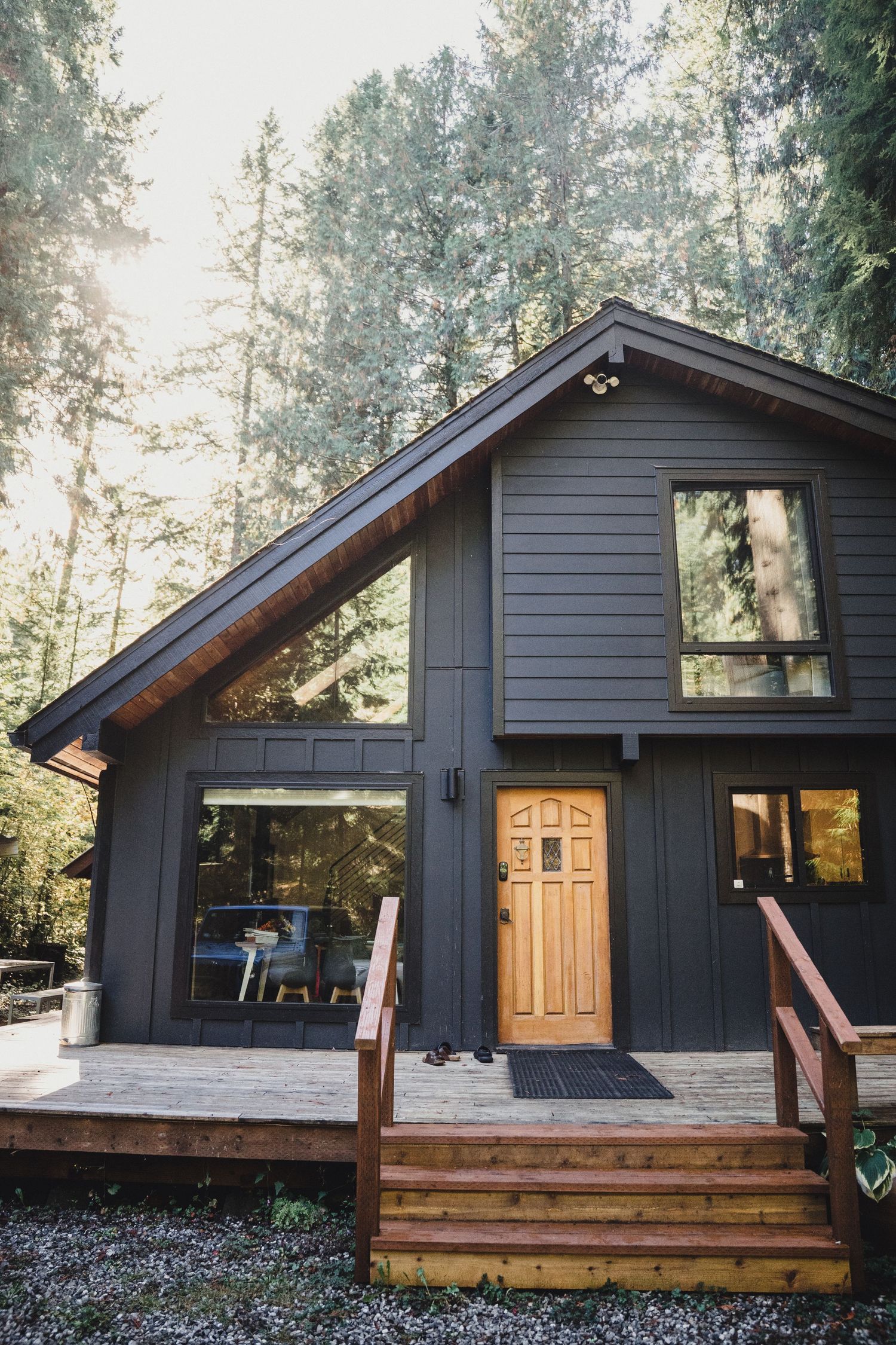 Dark gray A-frame cabin with a warm wooden front door, large windows, and a cedar deck with stairs, nestled among tall evergreen trees.