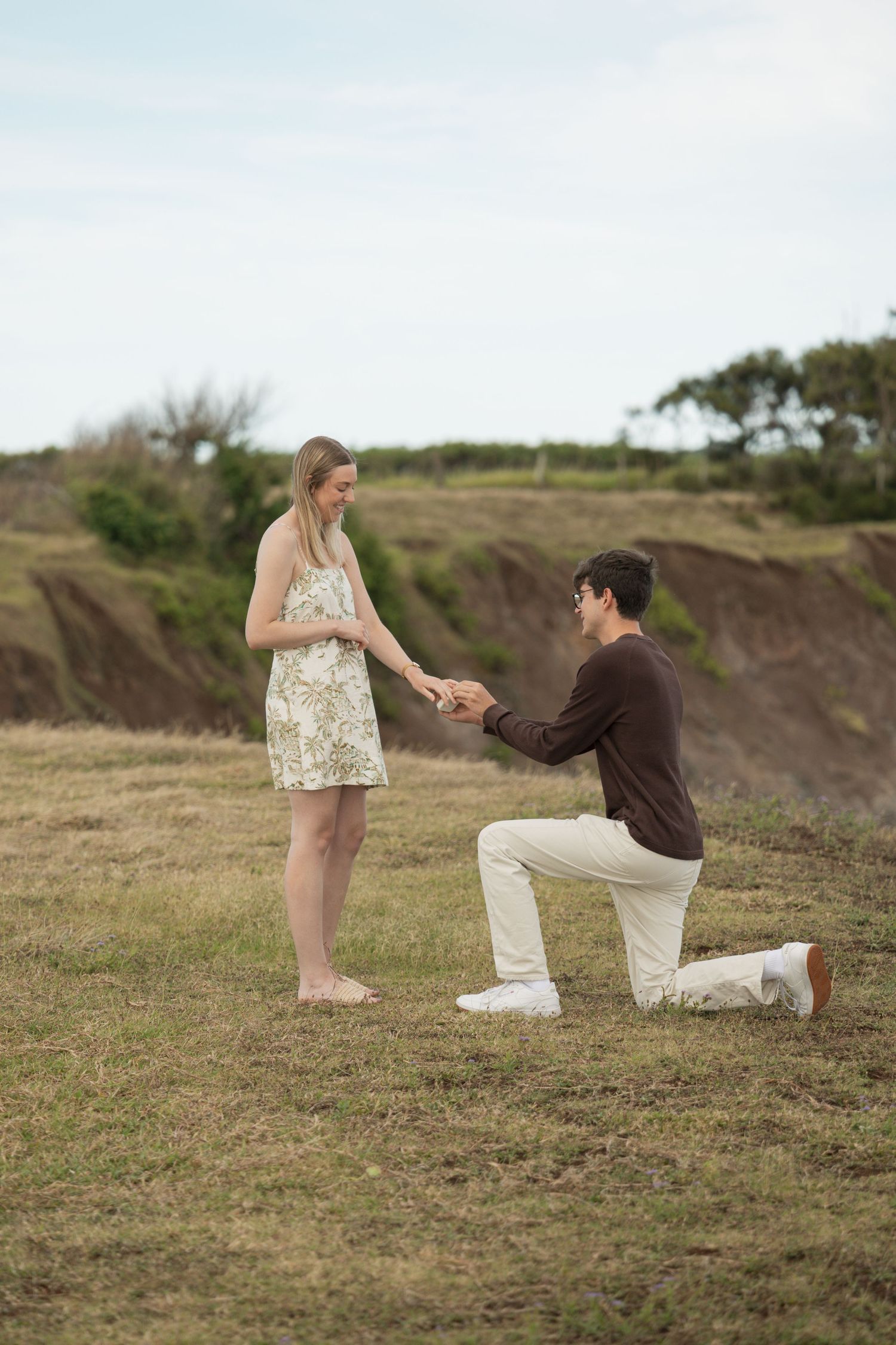 Man kneeling on one knee proposing to woman in floral dress outdoors on grassy hillside.