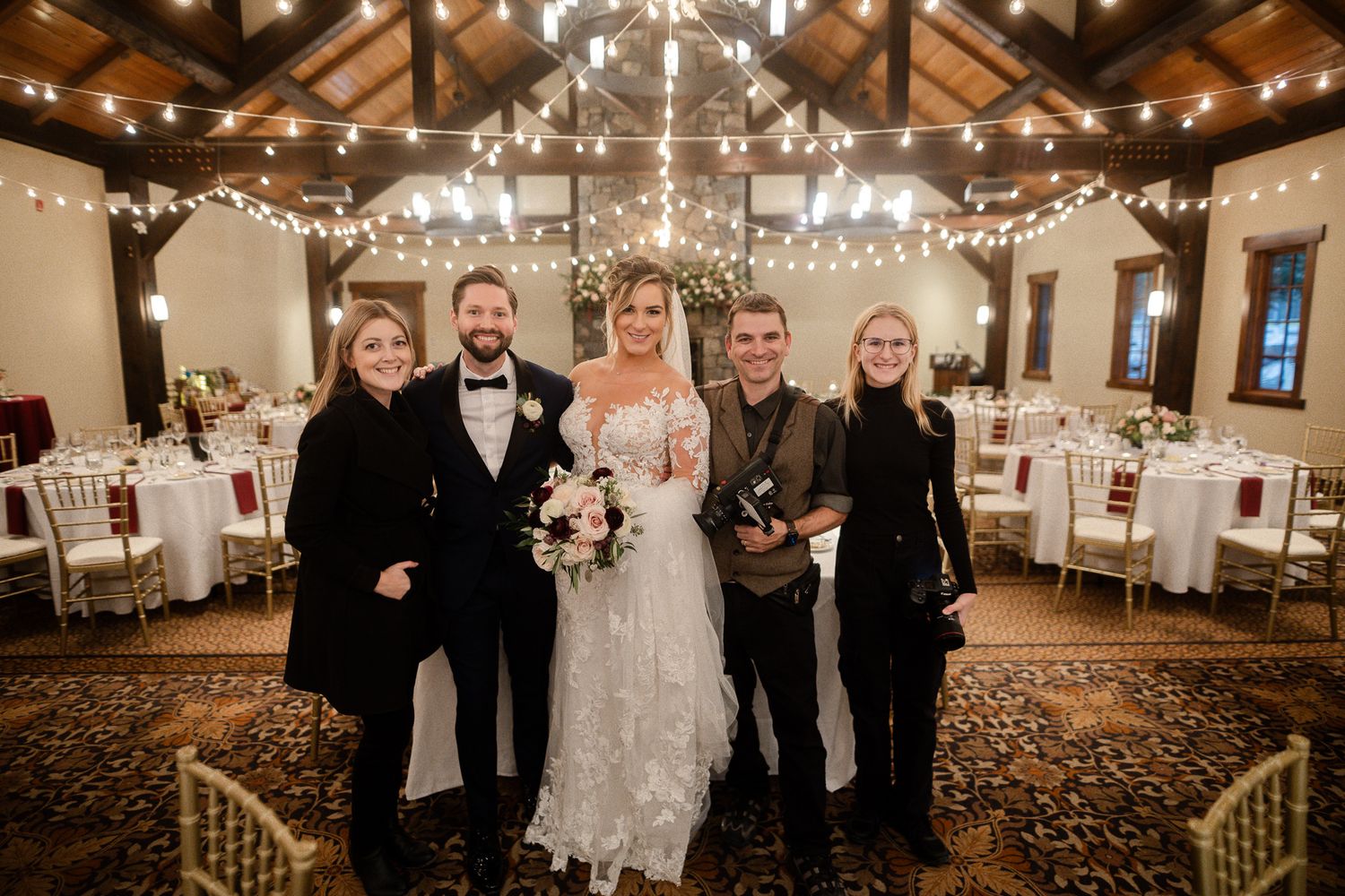 Bride and groom pose with three guests inside a rustic wedding venue with wood beams, string lights, and elegantly set tables.