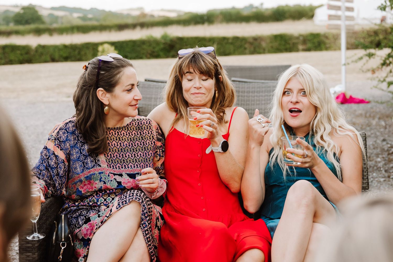 Candid wedding Three smiling women in colourful dresses sit close together outdoors, laughing and holding drinks at Cott Farm Barn Somerset