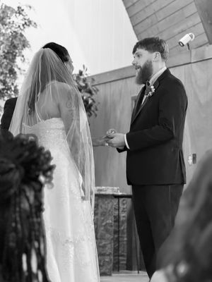A bearded groom in a dark suit exchanges rings with his veiled bride during a black and white wedding ceremony.