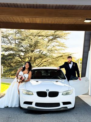 Bride in a white gown holding a bouquet and groom in a black suit pose beside a white BMW under a covered driveway.