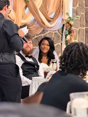 A smiling bride in a pearl necklace sits beside her groom in a bow tie as a guest gives a toast at the reception.