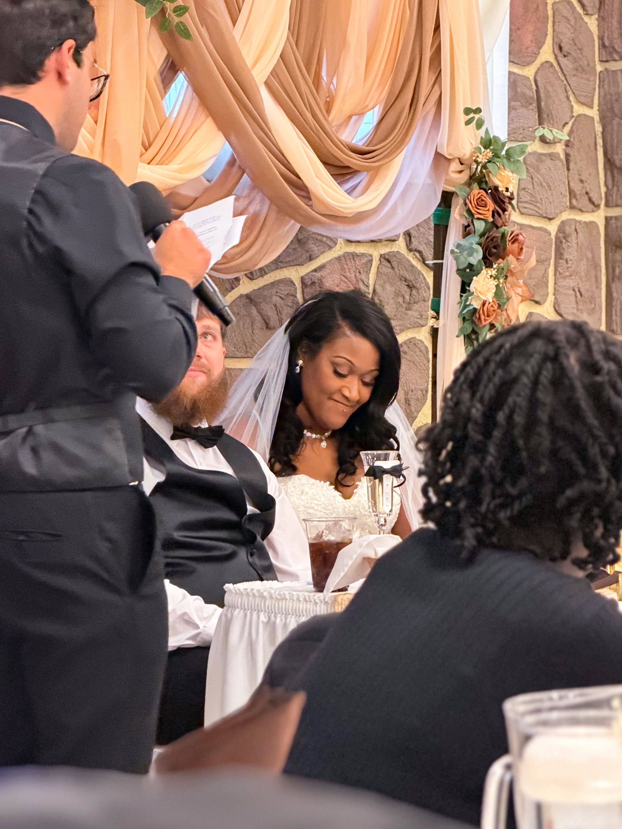 Smiling bride in a veil sits beside a bearded groom in a bow tie as a man speaks into a microphone at a wedding reception.