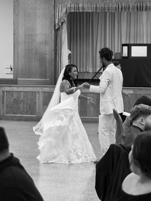 Black and white photo of a bride in a lace gown and a groom in a light suit joyfully dancing as wedding guests watch.