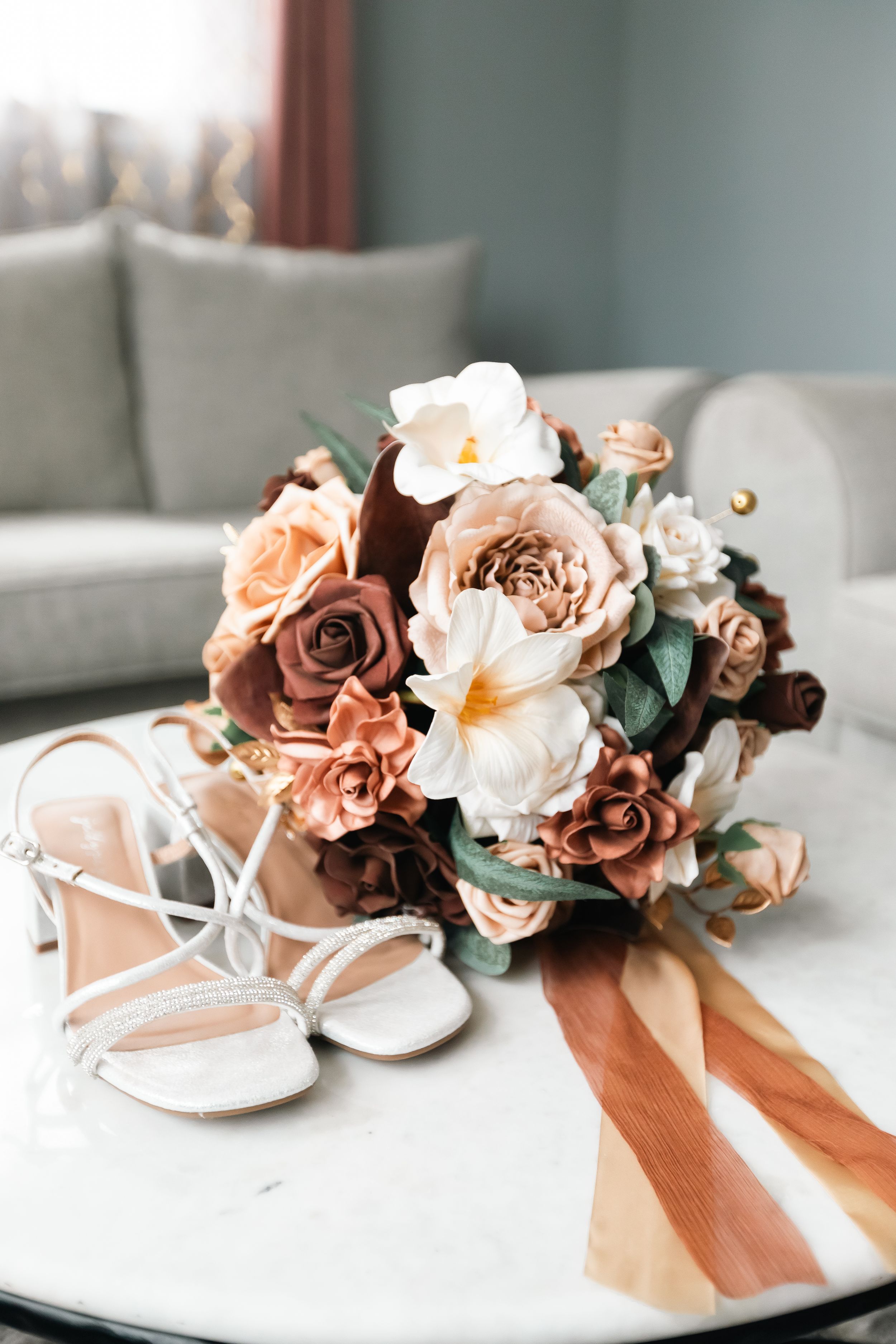 Bridal bouquet of terracotta roses, cream magnolias and greenery with rust ribbon beside white strappy heels on a marble table.