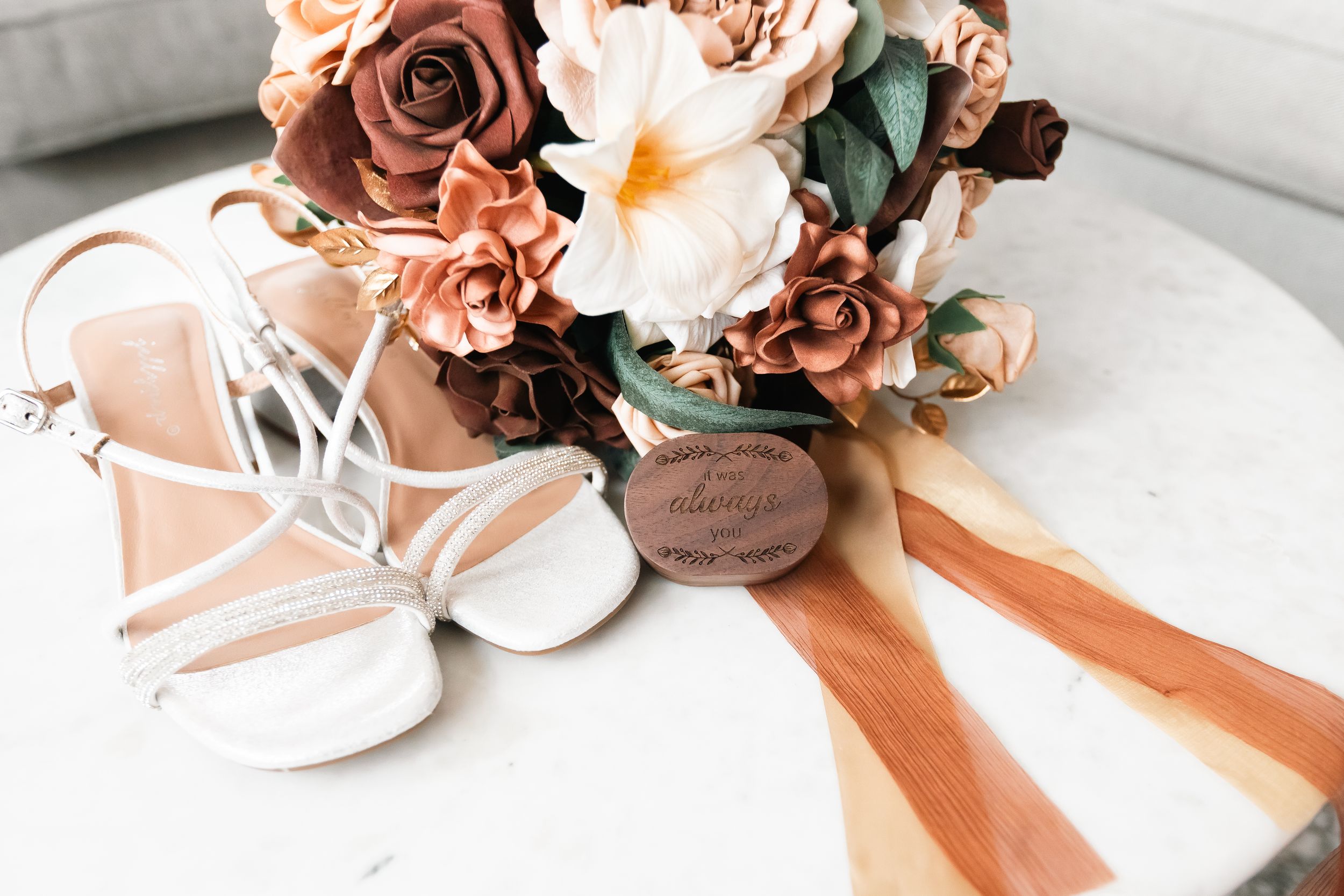 White strappy bridal sandals beside a bouquet of terracotta and cream roses with orange silk ribbons and a wooden charm on a marble table.