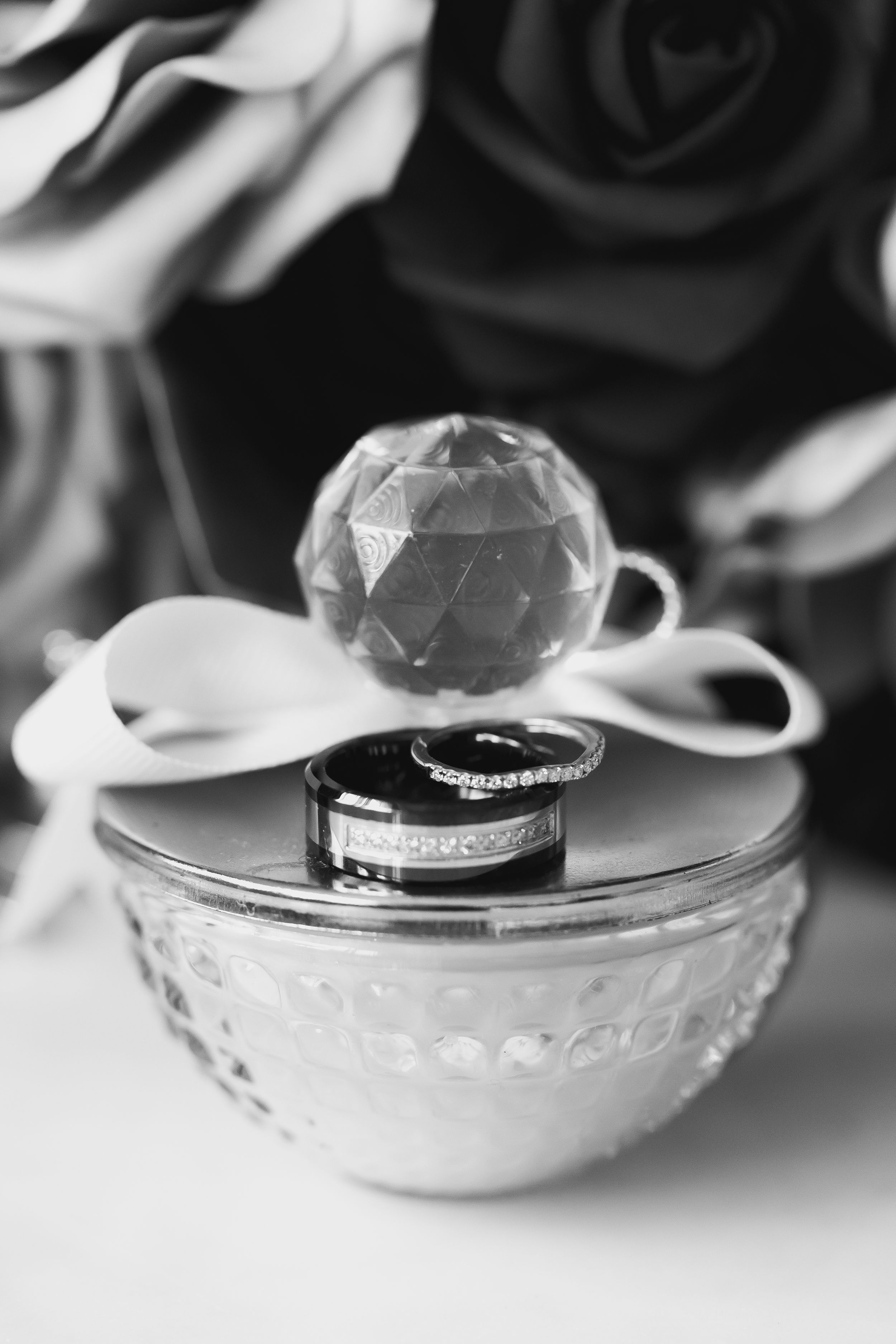 Black and white photo of two wedding rings resting on a glass perfume bottle with a faceted crystal stopper, bow, and roses behind.