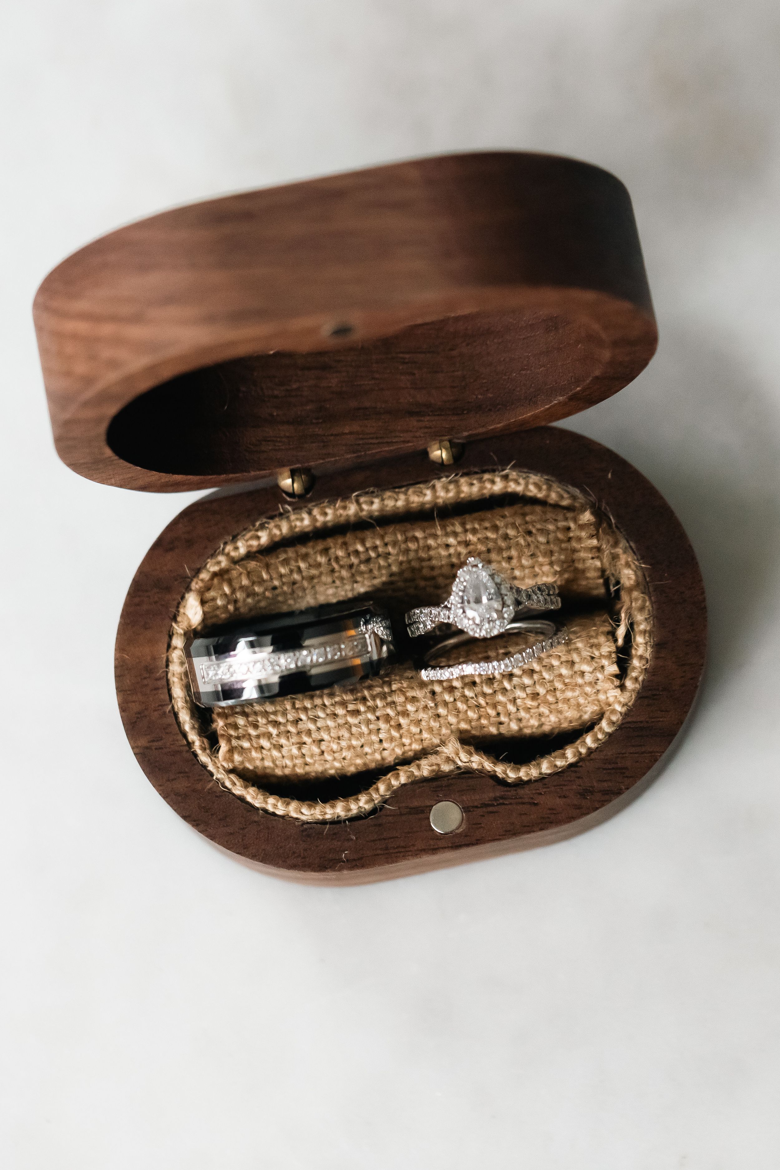 Wedding bands and a diamond halo engagement ring resting in an open oval walnut ring box lined with burlap.