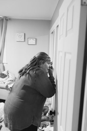 Black and white photo of a woman in a button-up shirt leaning toward a door while applying makeup in a bedroom.