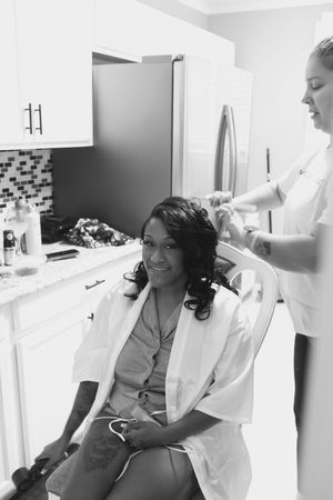 A smiling bride in a white satin robe sits in a kitchen chair while a stylist curls her hair on her wedding day.