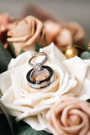 Diamond engagement ring and black wedding band resting inside a white rose surrounded by blush and peach roses in a wedding bouquet.
