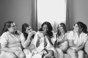 Black and white photo of five bridesmaids in matching robes laughing together while holding champagne glasses on a couch before the wedding.