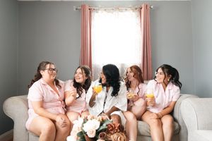 Bride in white robe sits with four bridesmaids in matching pink pajamas, holding mimosas and laughing on a gray sofa with a bouquet.