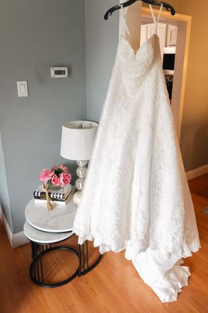 A white lace wedding gown with a sweetheart neckline hangs beside a marble side table with pink peonies and a lamp.