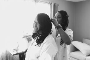 Black and white photo of a bridesmaid in a white robe styling the bride's long curly hair beside a sunlit window.