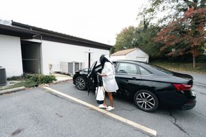 A bride in a white robe stands beside an open door of a black sedan in a parking lot, ready to leave for her wedding.