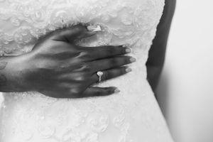 Black and white close-up of a bride's hand with a diamond engagement ring resting on her lace wedding dress.