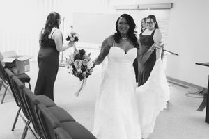 Smiling bride in a white wedding gown holds a bouquet while bridesmaids in matching dresses stand nearby in a bright ceremony room.