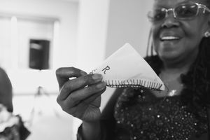 Smiling woman in glasses and a sparkly top holds up a folded paper triangle with the letter R and stitched edge in a black and white photo.