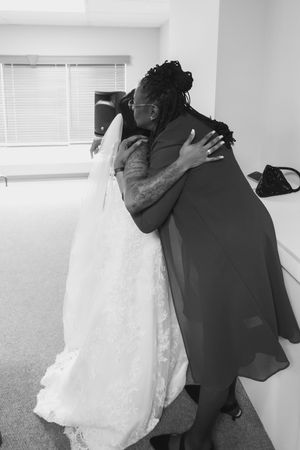 Black and white photo of a bride in a lace wedding gown and veil embracing a woman in a flowing dark dress in a bright room.