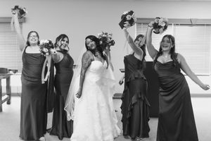 Black and white photo of a bride in a lace gown posing joyfully with four bridesmaids in long dark dresses holding bouquets.