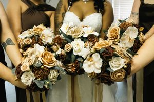 Bride in a white gown stands among bridesmaids holding matching bouquets of cream, ivory and brown roses and white magnolia-style blooms.