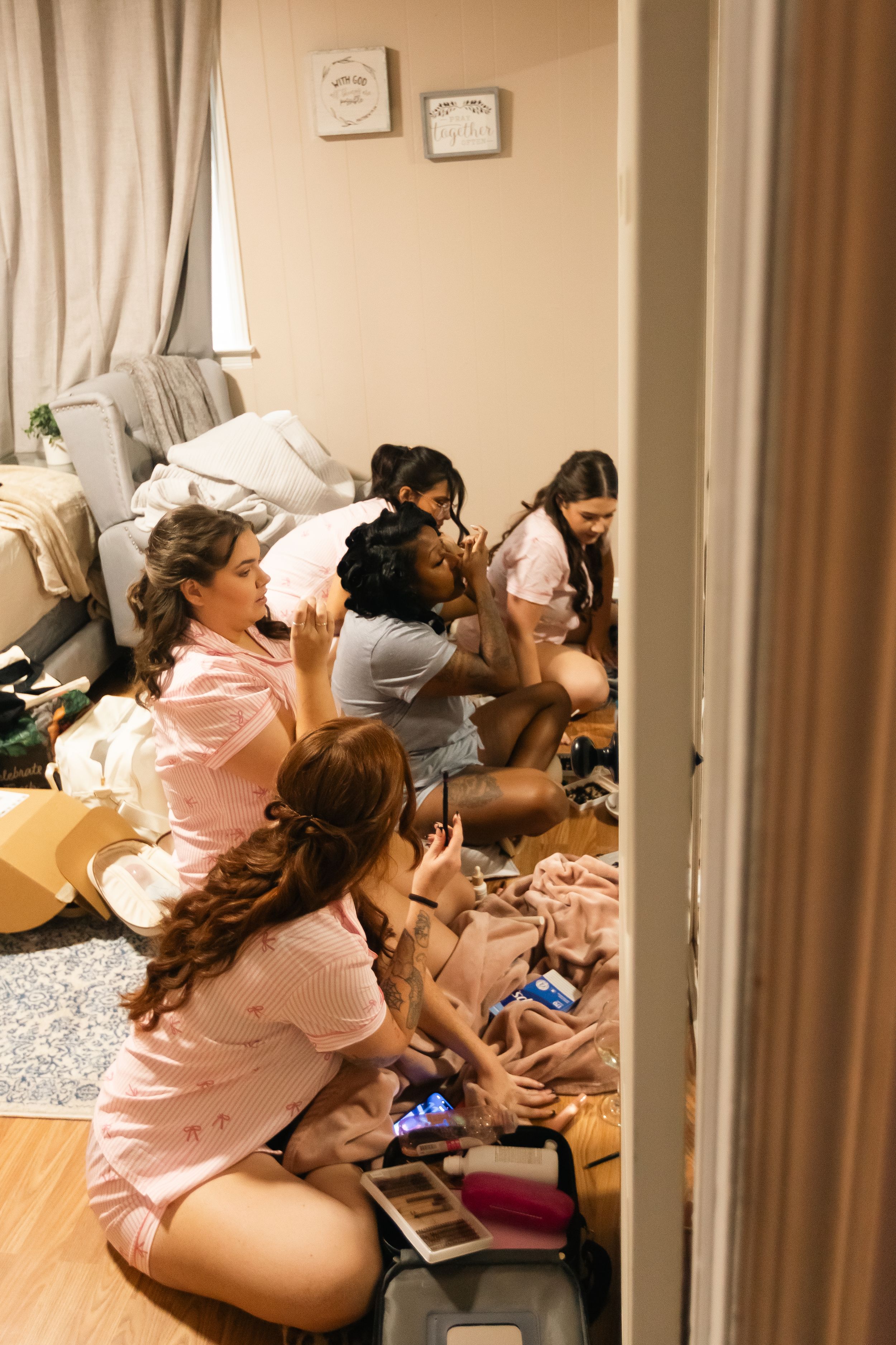 Bridesmaids in matching pink pajamas sit on the floor getting ready together on the wedding morning with makeup and hair supplies.