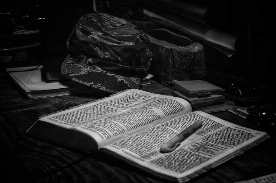 Black and white photo of an open Bible on a table with a pen and dark objects in the background.