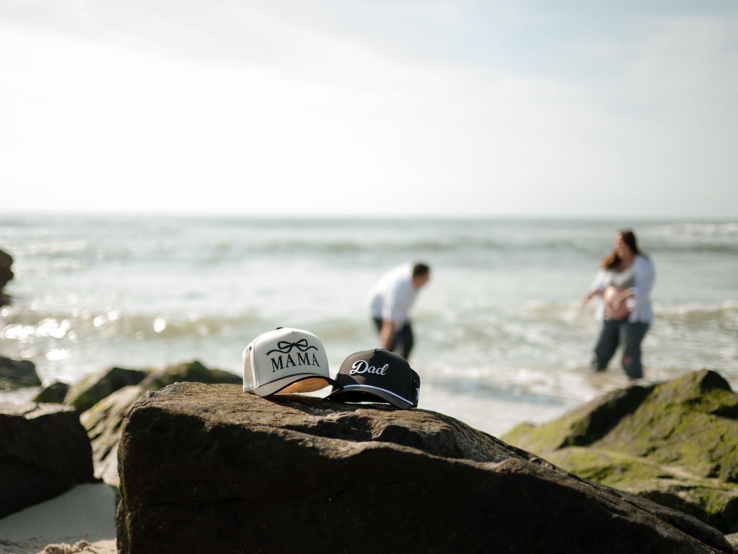 Mama and Dad caps sit on a seaside rock while a blurred expecting couple plays in the ocean waves in the background.