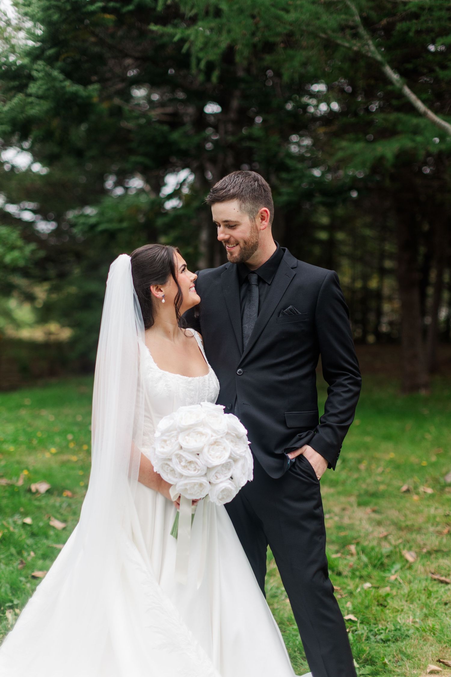 Smiling bride in a white gown, veil and white rose bouquet embraces her groom in a black suit on a green lawn near evergreen trees.