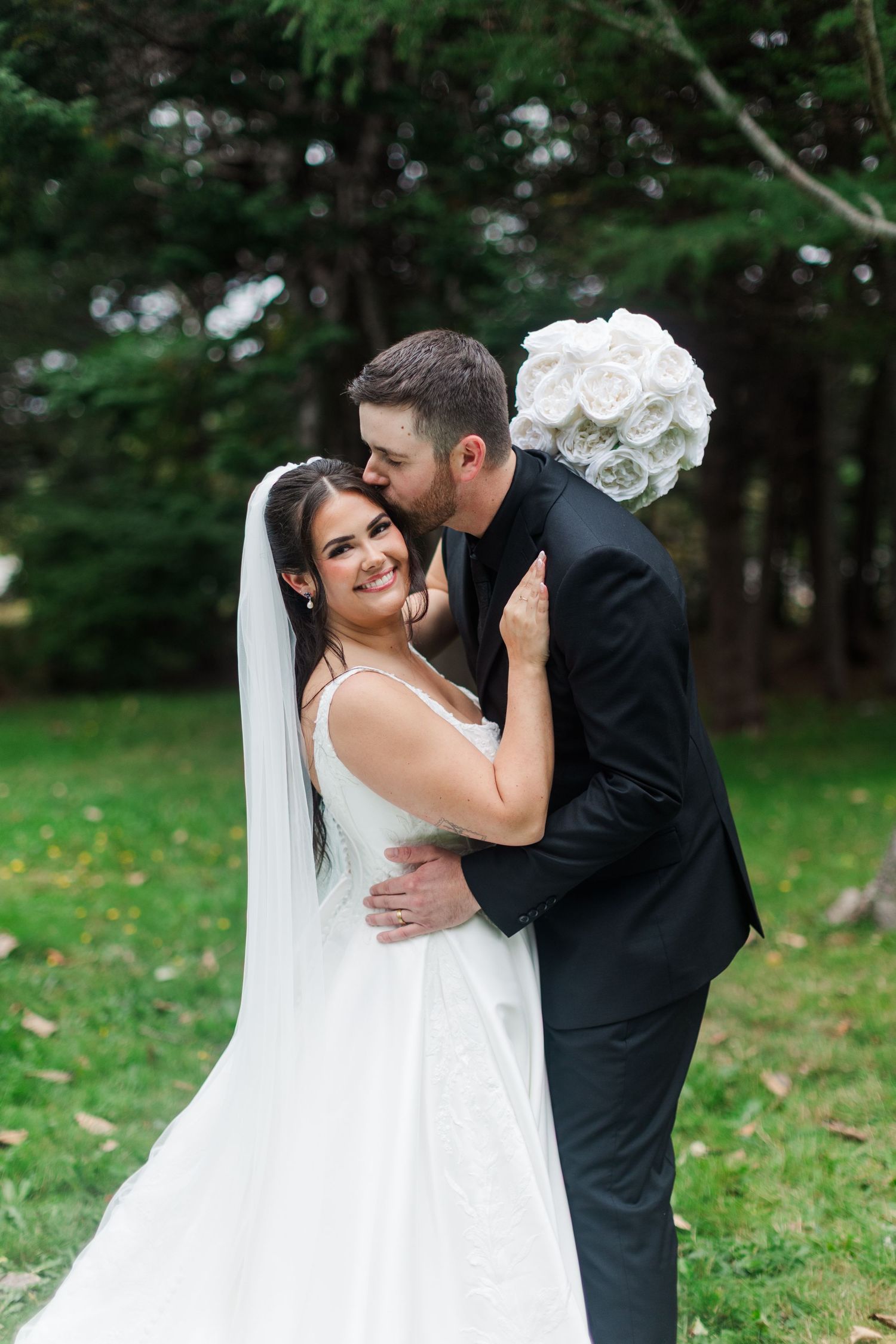Smiling bride in a white gown and veil embraces her groom in a black suit as he kisses her cheek, holding a white bouquet outdoors.
