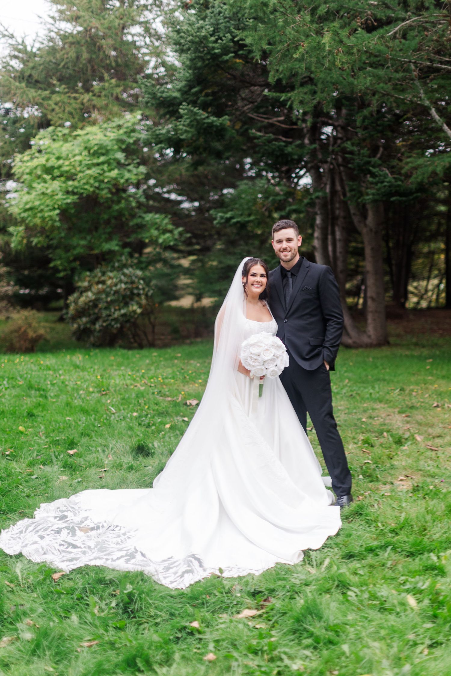 Bride in a white gown with a long lace-trimmed train holds a bouquet beside her groom in a dark suit on a green lawn with trees.