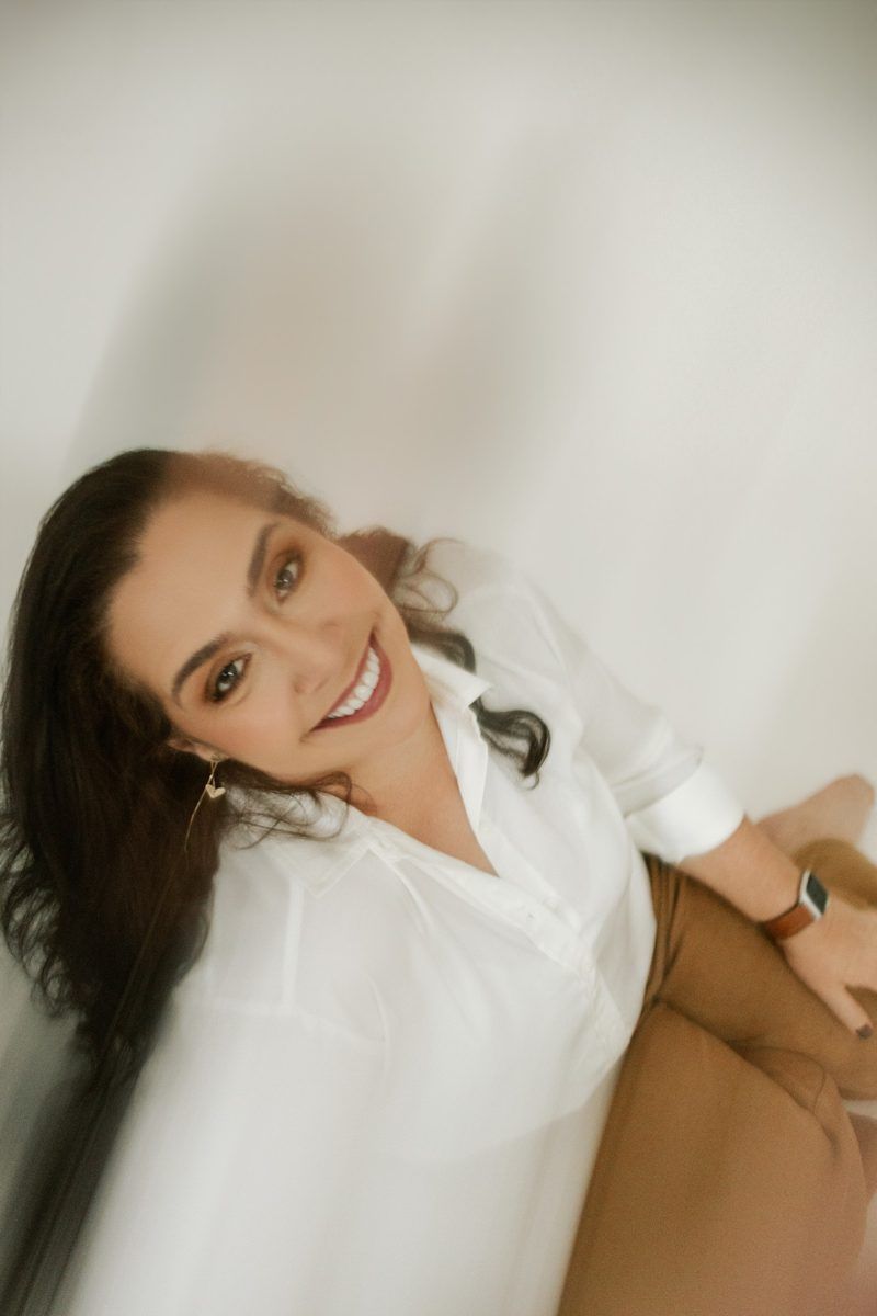 Smiling woman with long dark hair in a white button-up shirt and tan pants sits on a white background, wearing earrings and a smartwatch.