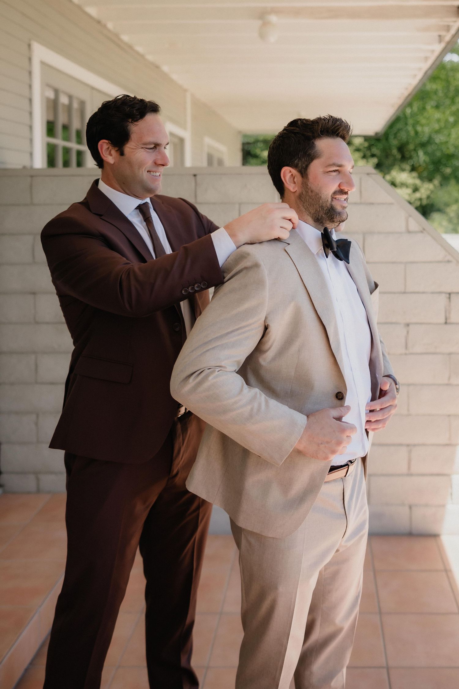 Groomsman helping groom in beige suit adjust his jacket before wedding ceremony.