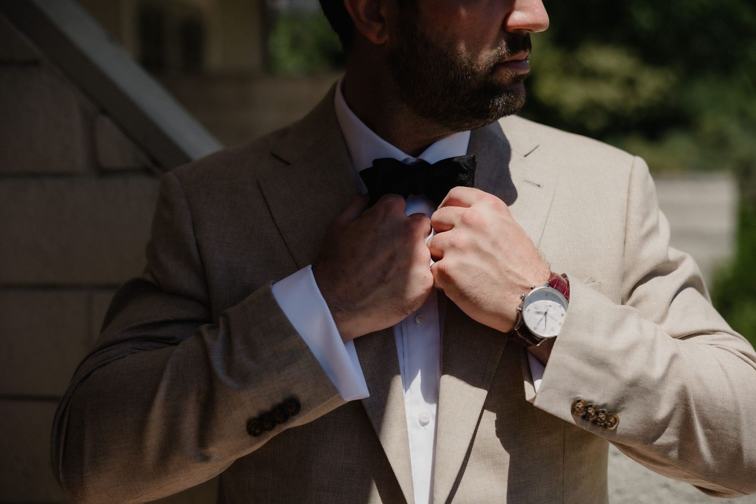 Man in beige suit adjusting black bow tie, wearing a watch, outdoors in natural light.