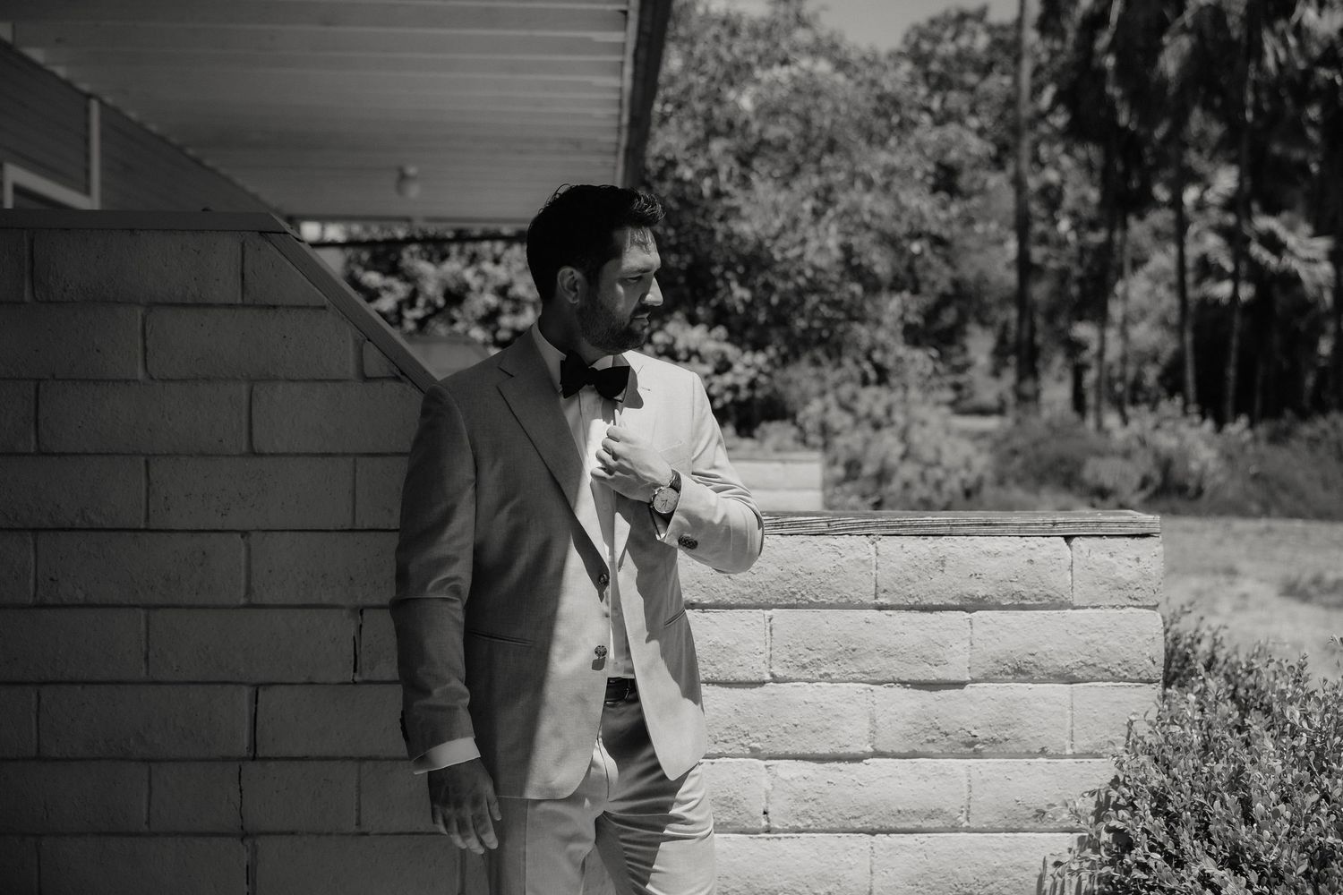 Man in suit reading paper outdoors leaning against brick wall, black and white wedding photography.