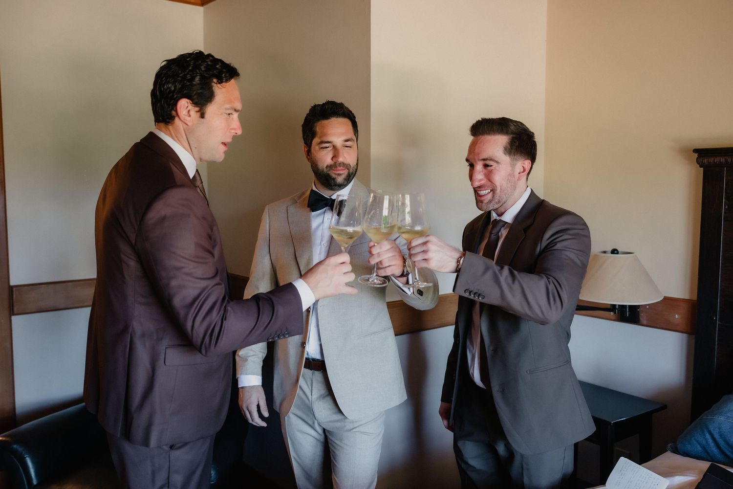 Three groomsmen in suits toasting with champagne glasses during wedding day preparations.