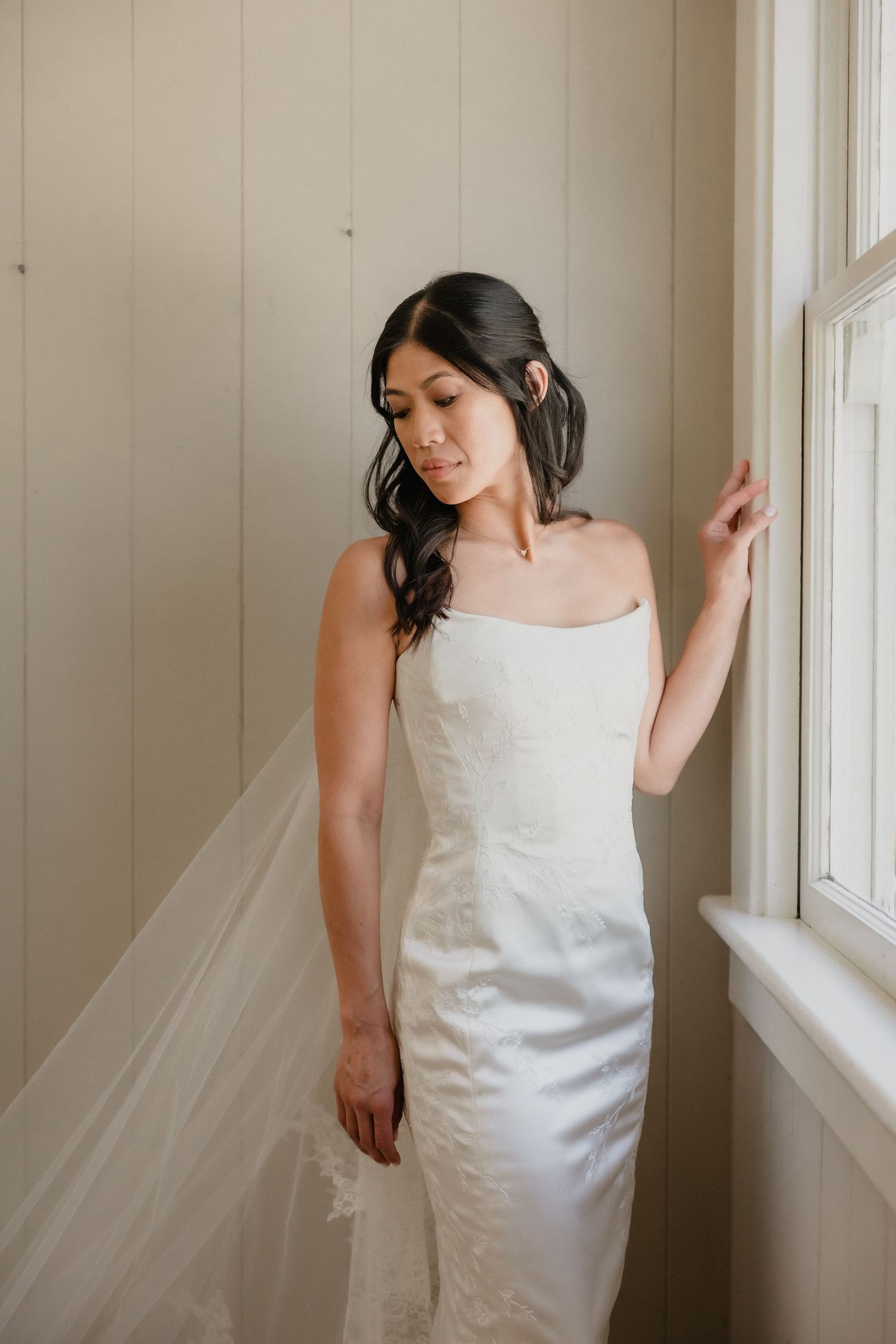 Bride in strapless white satin gown with lace cathedral veil standing by window in soft light.