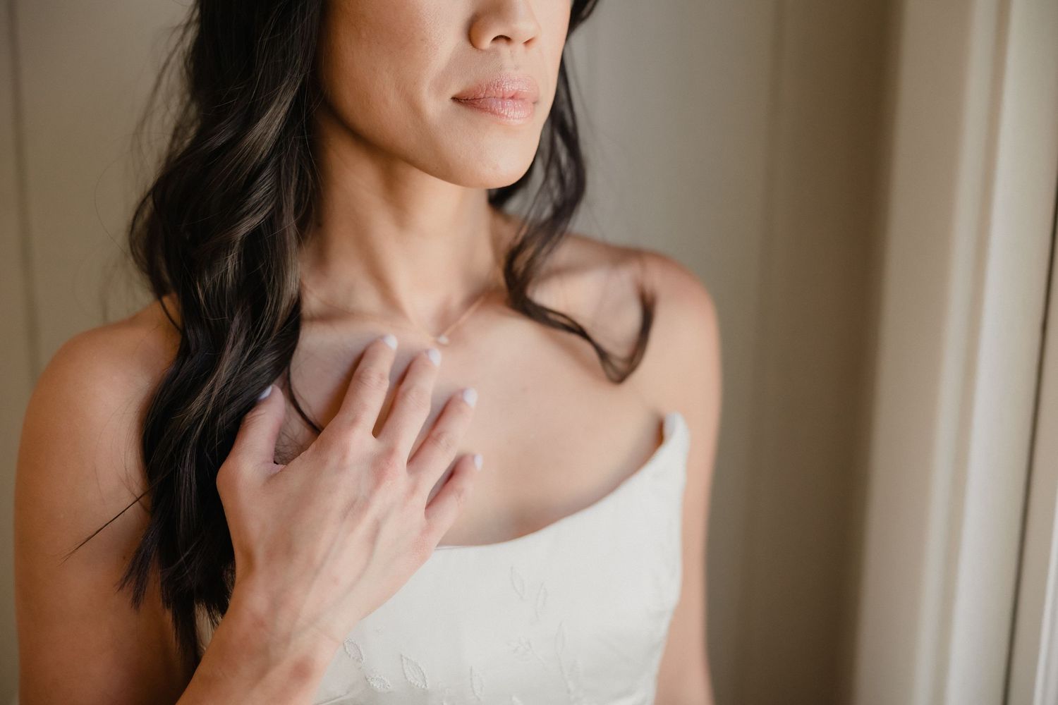 Bride in white strapless gown touching her collarbone, dark wavy hair, elegant bridal portrait.
