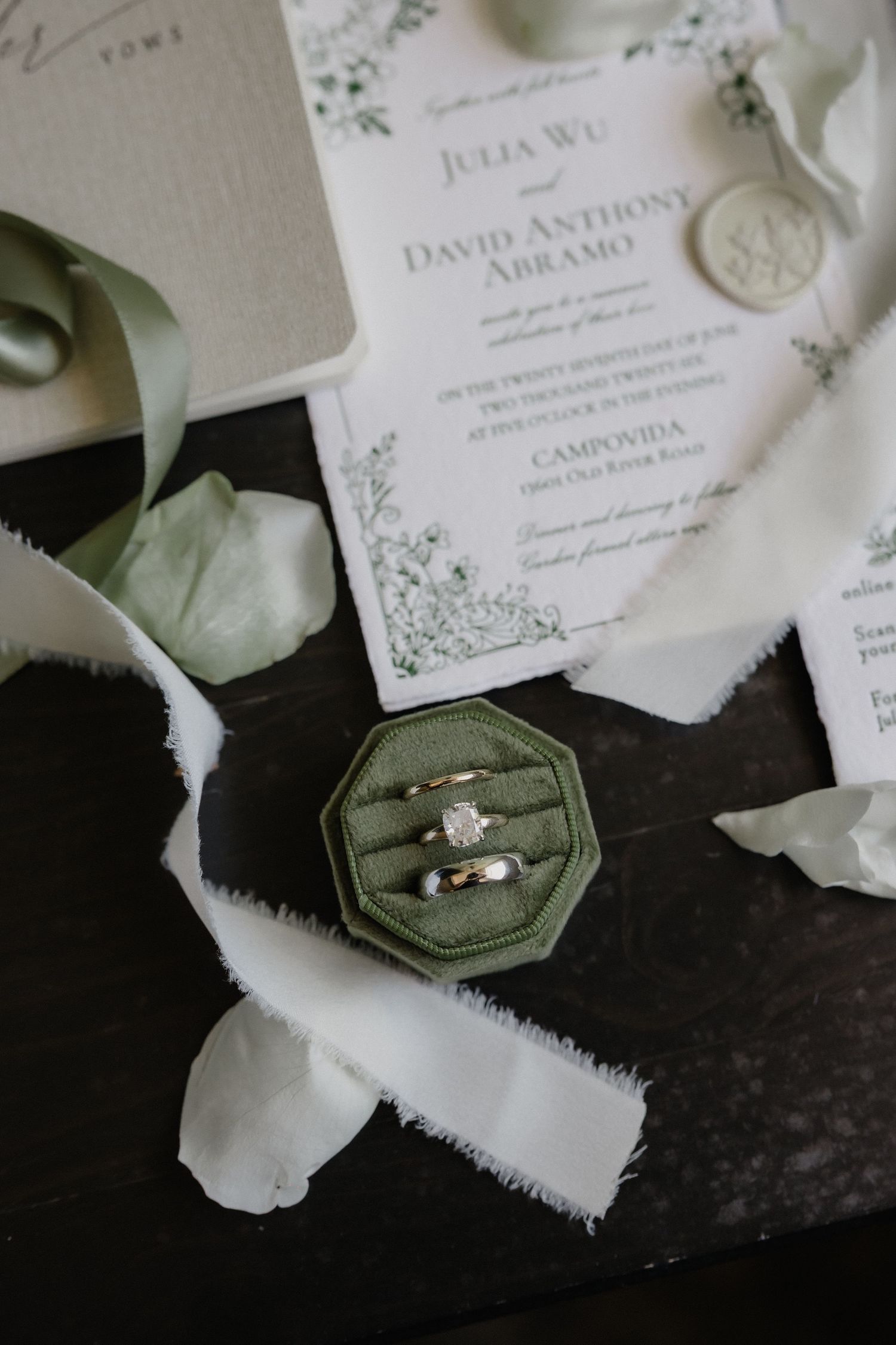 Wedding rings in a green velvet box beside floral invitation for Julia Wu and David Anthony Abramo.